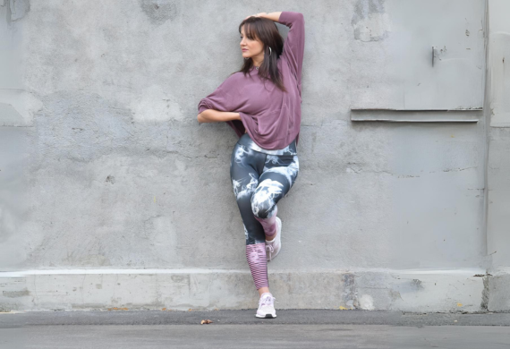 Eine Person in Sportkleidung lehnt an einer grauen Wand, schaut weg, hat ein Knie angewinkelt und den Fuß an der Wand.